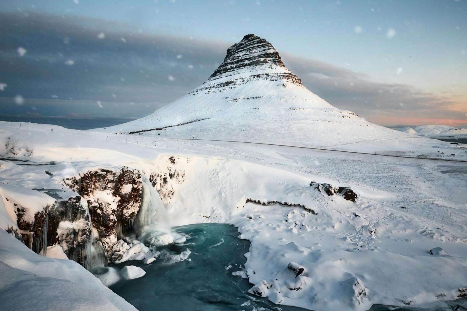 Snow-covered Kirkjufell rises behind icy waterfalls and a turquoise pool