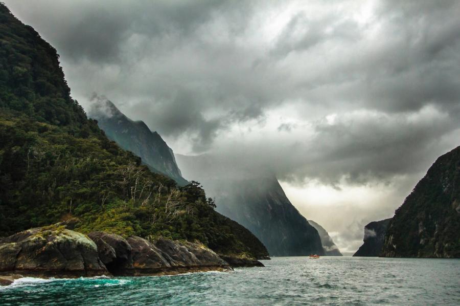 Clouds hang over steep forested mountains and a boat in Milford Sound