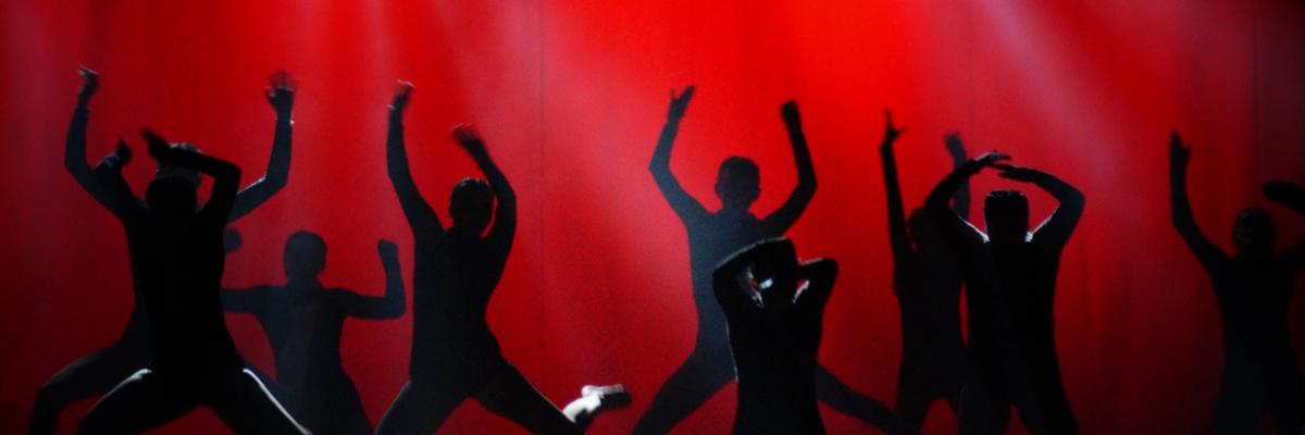Silhouetted dancers performing on stage under red theater lighting during a live arts performance.