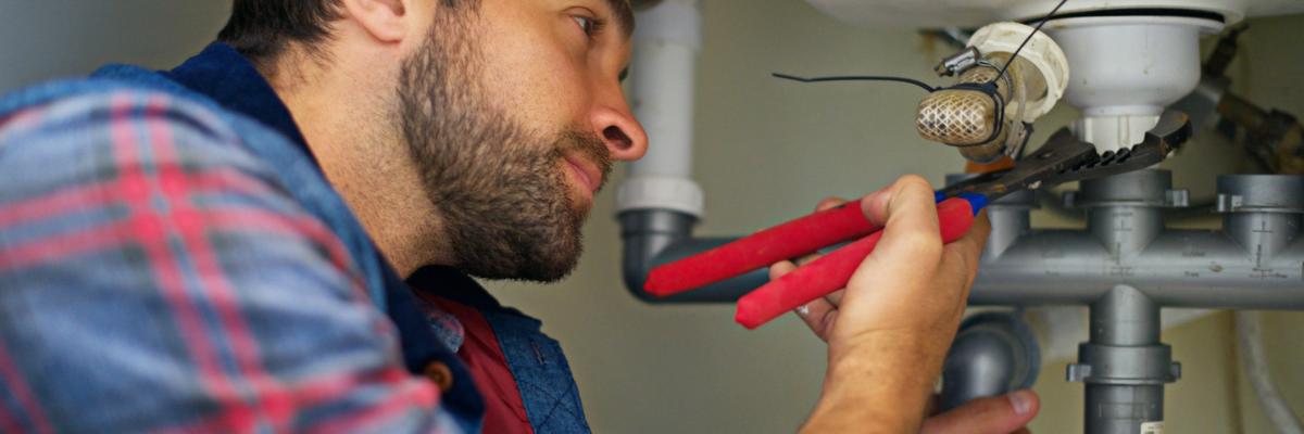 Professional plumber using pliers to repair a kitchen sink drain pipe and P-trap beneath a cabinet in a residential home.