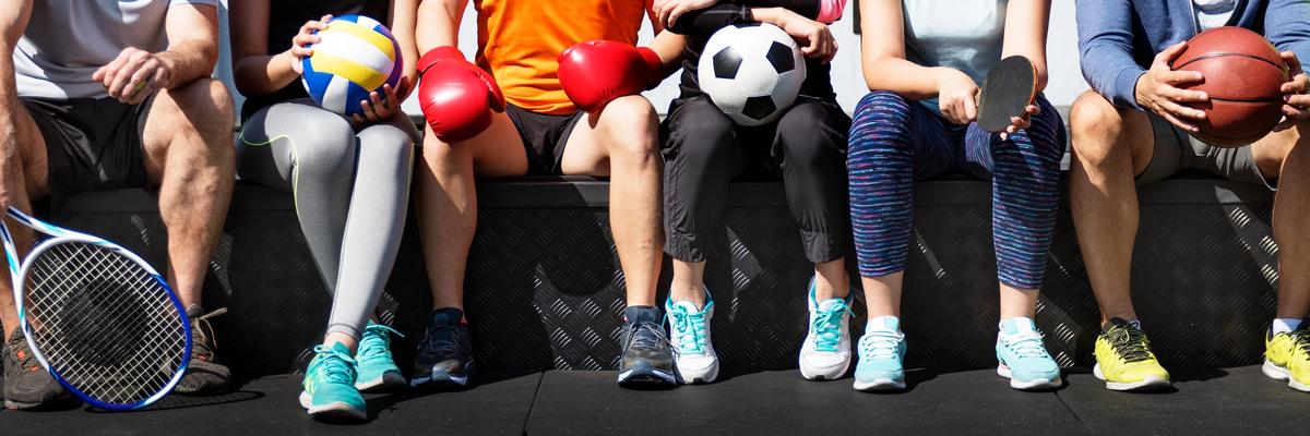 Group of athletes sitting together holding sports equipment including soccer ball, basketball, volleyball, tennis racket, and boxing gloves.