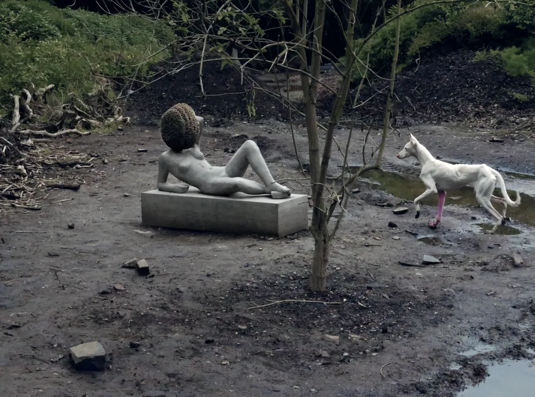 The reclining sculpture with a beehive head and a white dog in Untilled