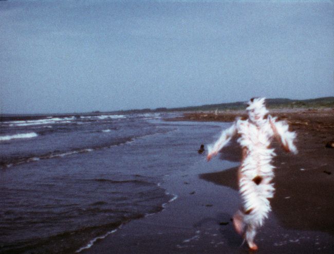 Covered in white feathers, Mendieta runs towards the camera along a beach