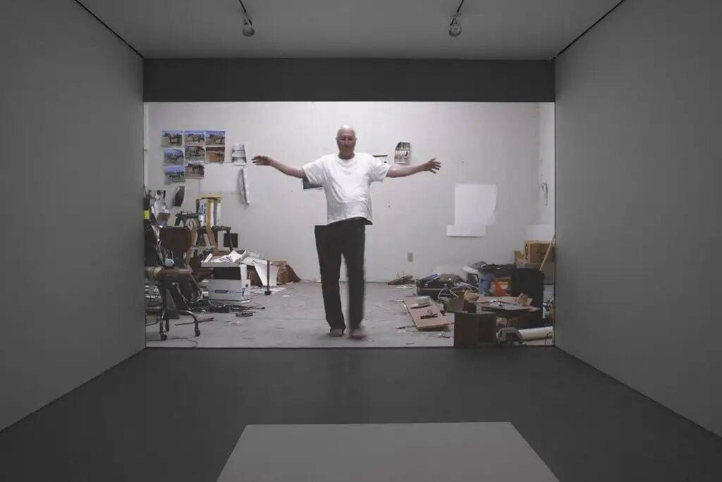A darkened art gallery with a video of the artist Bruce Nauman standing in his studio, arms outstretched