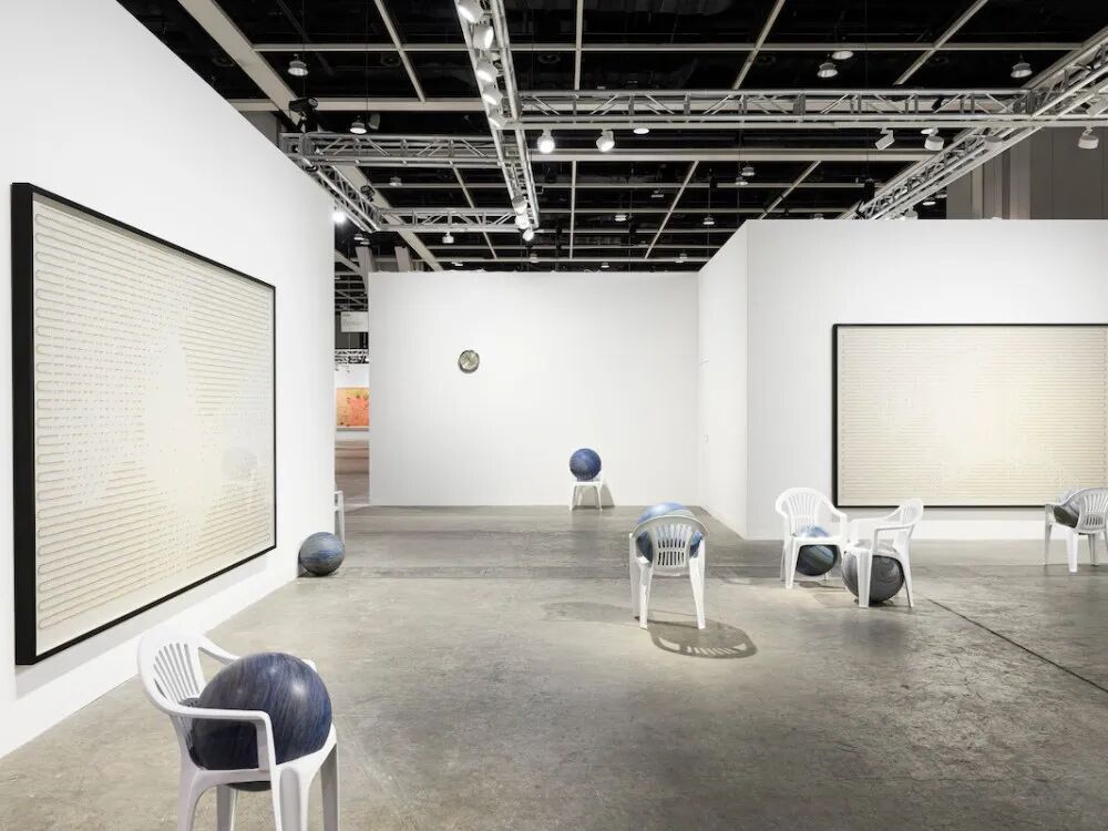 View of an art fair booth with white artworks on the walls and sculptures made of orbs cut into patio chairs.