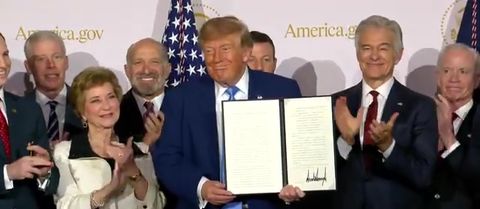 President Trump holds up the signed America.gov executive order at the Andrew W. Mellon Auditorium, September 29, 2026, with officials applauding (Official White House video still)