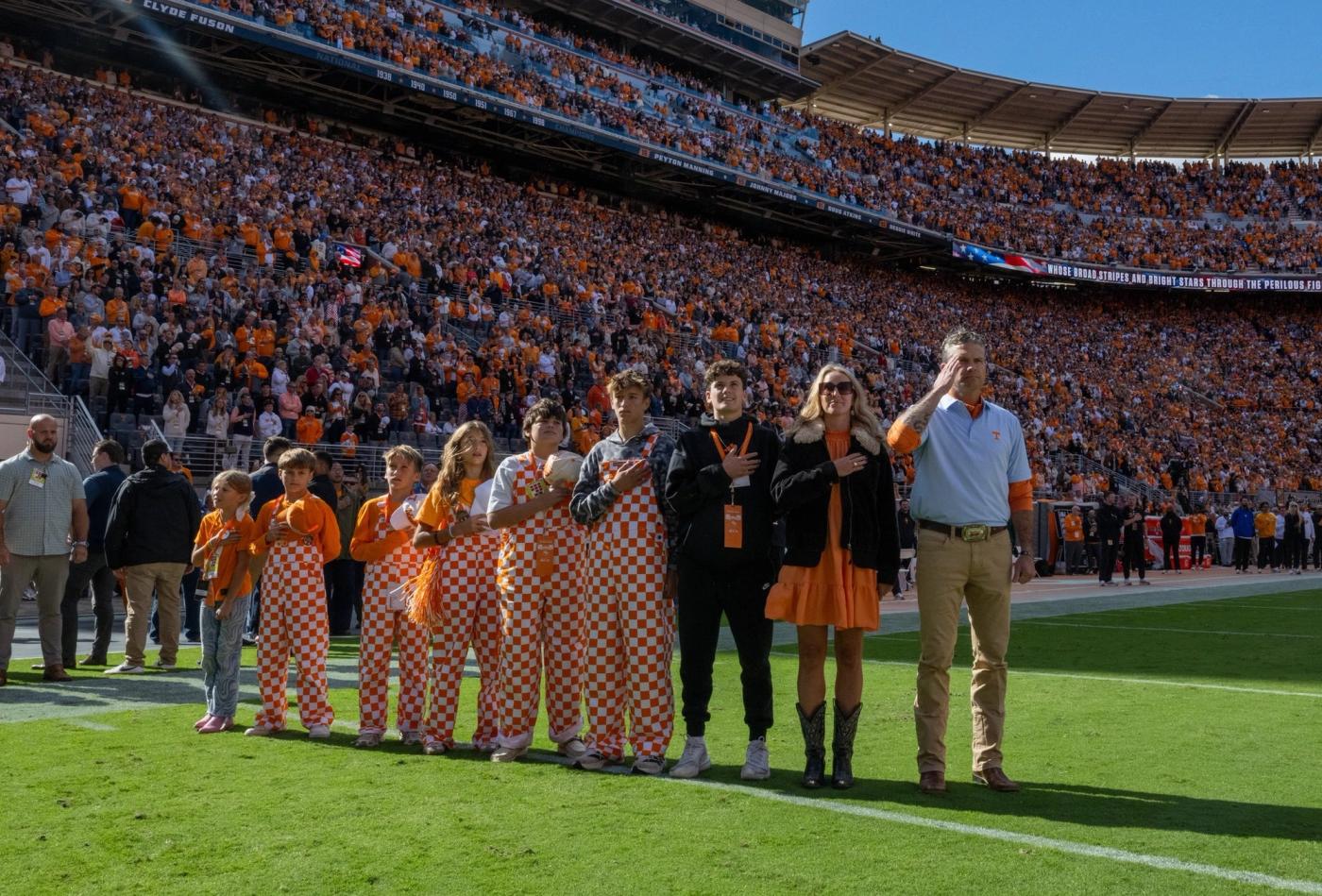 A man in a light blue polo salutes as children in orange-and-white checkerboard overalls and others stand with hands over their hearts on the field during the national anthem before a packed stadium crowd in Knoxville, Tennessee, Oct. 17, 2026. Secretary of War Pete Hegseth on X, Oct. 17, 2026: “For generations, the Volunteer State has raised its right hand and said, “SEND ME.””