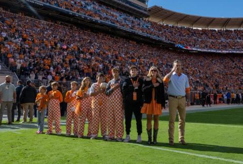 A man in a light blue polo salutes as children in orange-and-white checkerboard overalls and others stand with hands over their hearts on the field during the national anthem before a packed stadium crowd in Knoxville, Tennessee, Oct. 17, 2026. Secretary of War Pete Hegseth on X, Oct. 17, 2026: “For generations, the Volunteer State has raised its right hand and said, “SEND ME.””