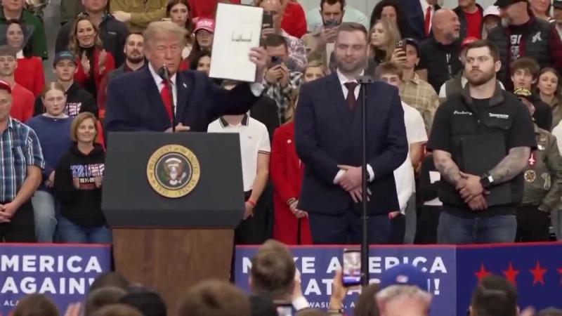 President Trump holds up a signed document at the podium as men stand beside him on stage at his rally in Wasilla, Alaska, October 13, 2026 (still from Rapid Response 47 video)