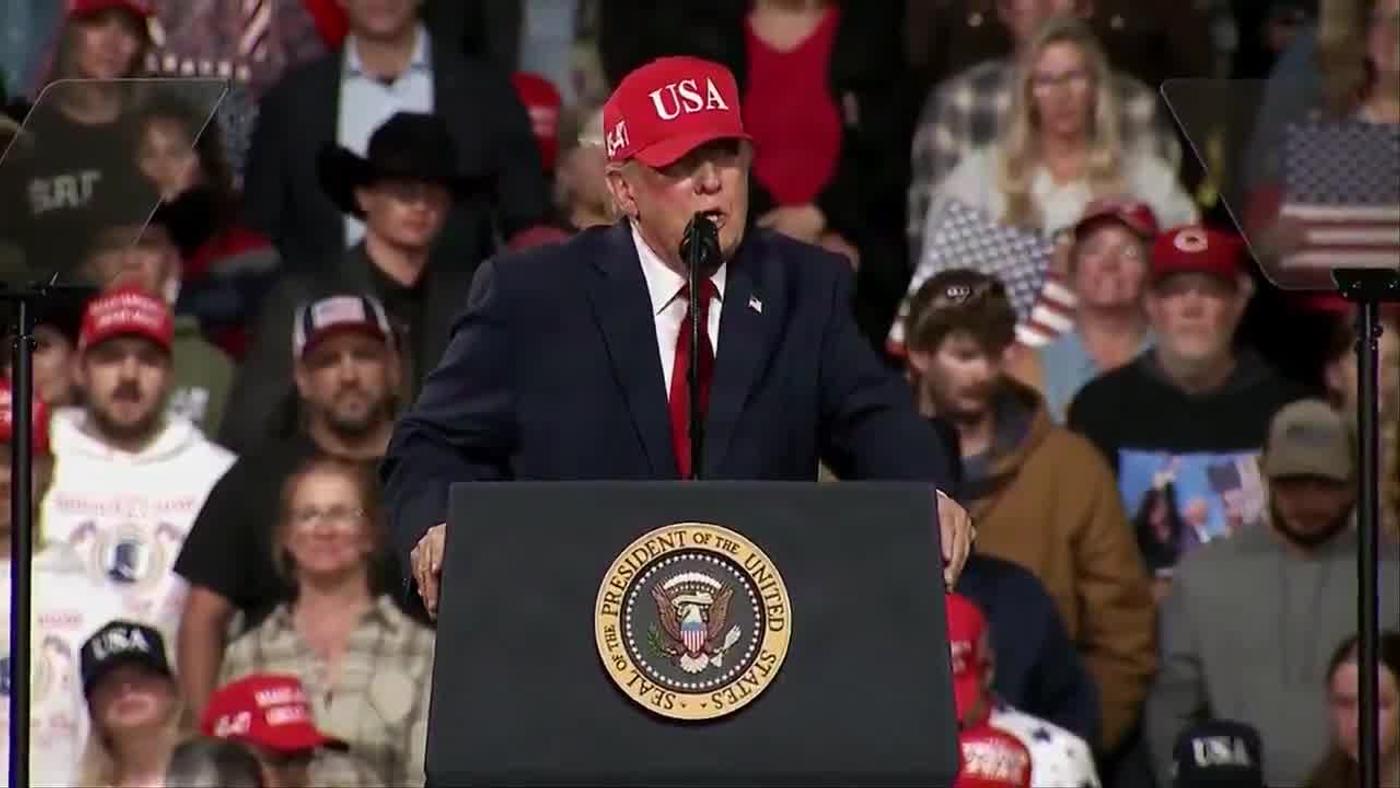 President Donald J. Trump, wearing a navy suit, red tie and red USA cap, speaks at a lectern bearing the presidential seal at his rally in Montana, October 14, 2026 (still from Rapid Response 47 video)