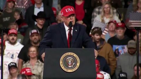 President Donald J. Trump, wearing a navy suit, red tie and red USA cap, speaks at a lectern bearing the presidential seal at his rally in Montana, October 14, 2026 (still from Rapid Response 47 video)