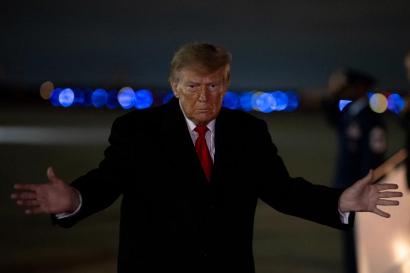 President Donald J. Trump, in a dark overcoat, gestures with both arms outstretched at night. (Official White House photo, Departure and Arrival gallery, October 10, 2026)