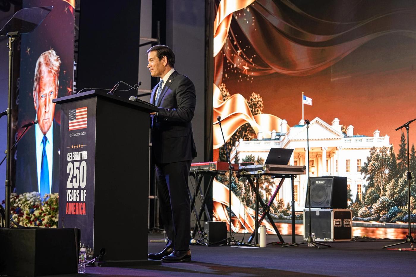 Secretary of State Marco Rubio speaks at a Freedom 250 reception in Mumbai, India, October 20, 2026, with an image of President Trump on screen (Official State Department photo by Freddie Everett / public domain)