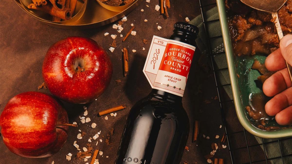 A bottle of 2026 Goose Island Bourbon County Apple Crisp Stout with a red label, surrounded by two whole red apples, scattered oats, cinnamon sticks, and a baking dish containing a freshly baked apple crisp being served with a spoon.