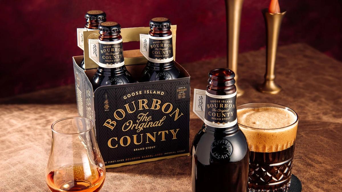 A four-pack of 2026 Goose Island Bourbon County The Original Brand Stout sitting on a textured brown surface. In the foreground, a dark glass of the stout with a thick tan head sits next to a single bottle, alongside a snifter glass containing a small pour of bourbon.