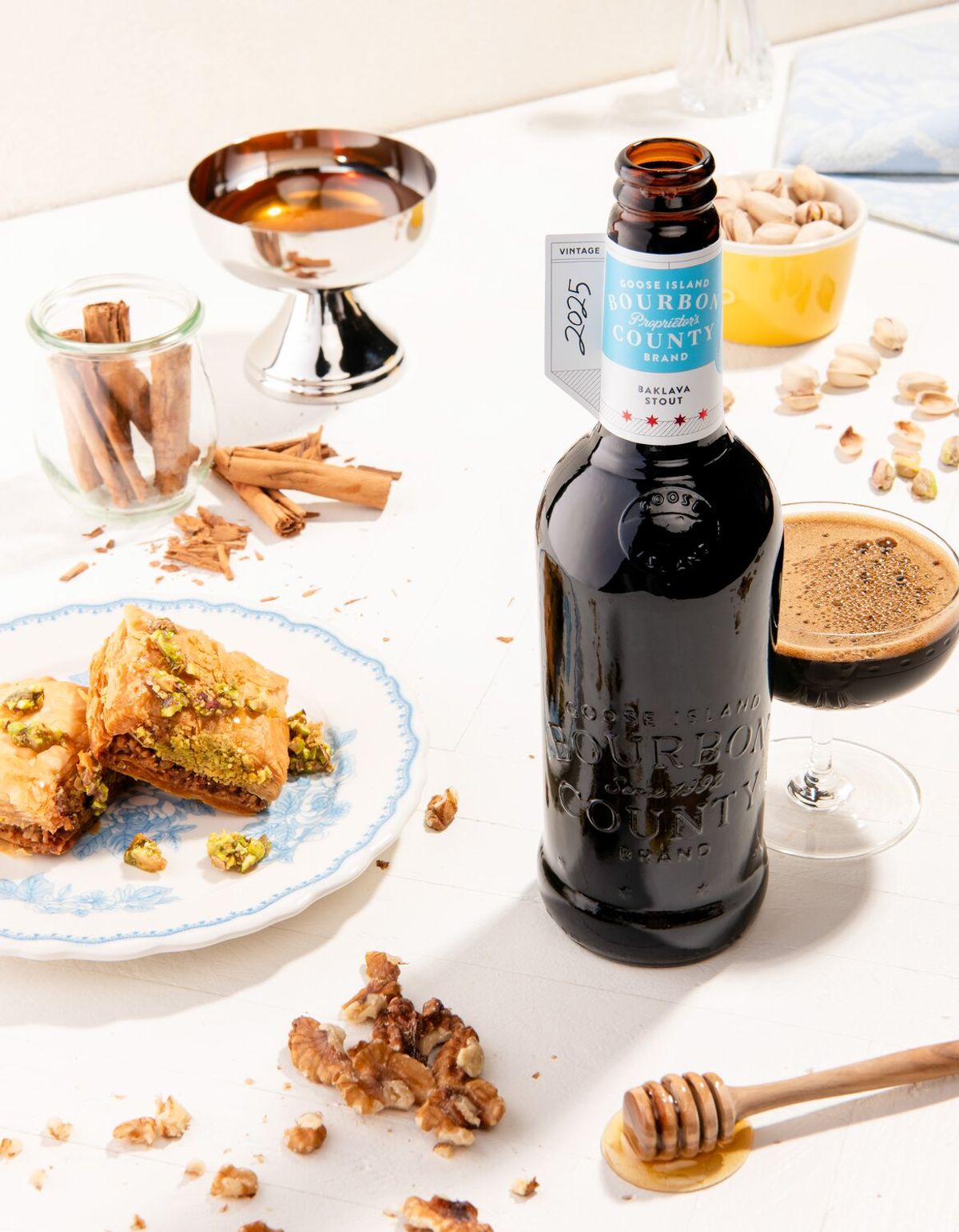 The 2025 blue-label Goose Island Proprietor's Baklava Stout bottle beside baklava, honey, walnuts and pistachios.