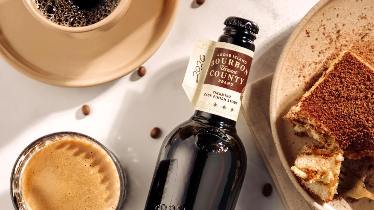Top-down view of a 2026 Goose Island Bourbon County Tiramisu Cask Finish Stout bottle. It is surrounded by scattered coffee beans, a cup of dark coffee, a glass of the stout, and a plate holding a slice of cocoa-dusted tiramisu.