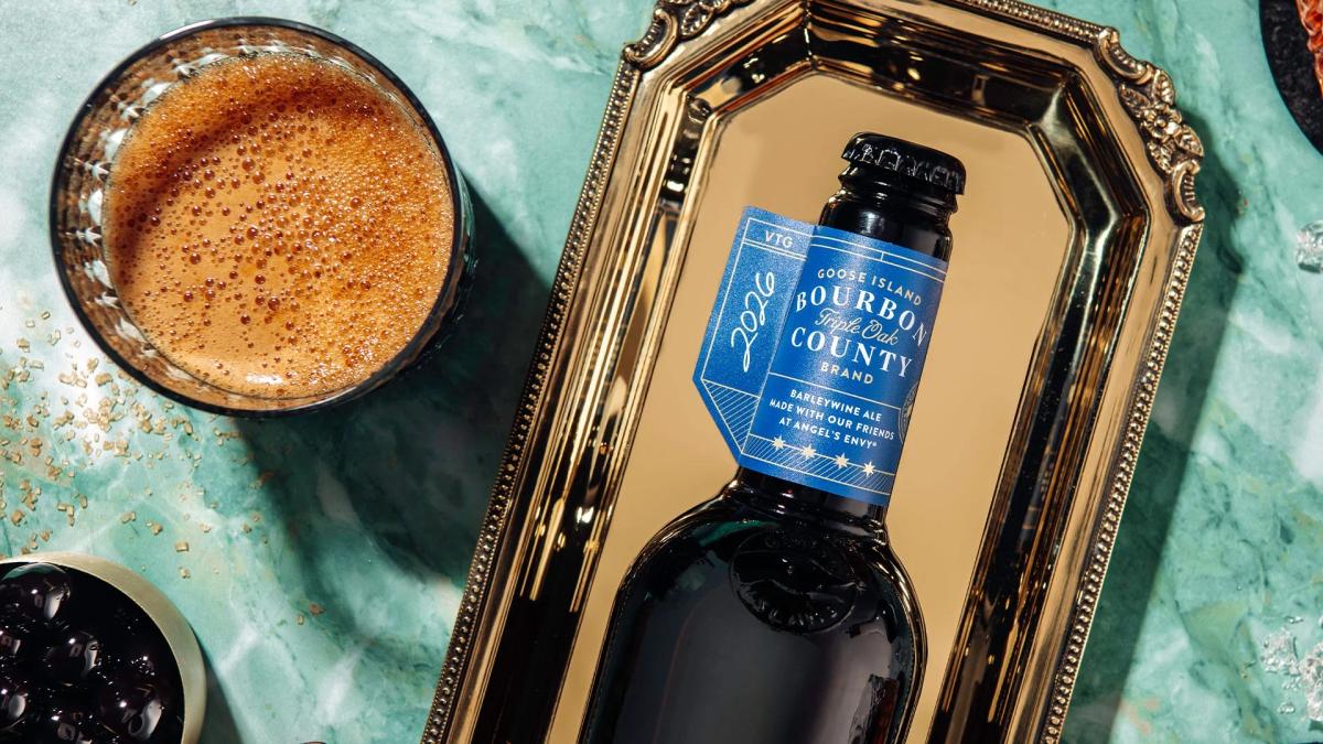 A bottle of 2026 Goose Island Bourbon County Triple Oak Barleywine Ale with a dark blue label lying elegantly on a gold serving tray over a green marble surface. A glass of the frothy ale and a small bowl of dark cherries sit nearby.