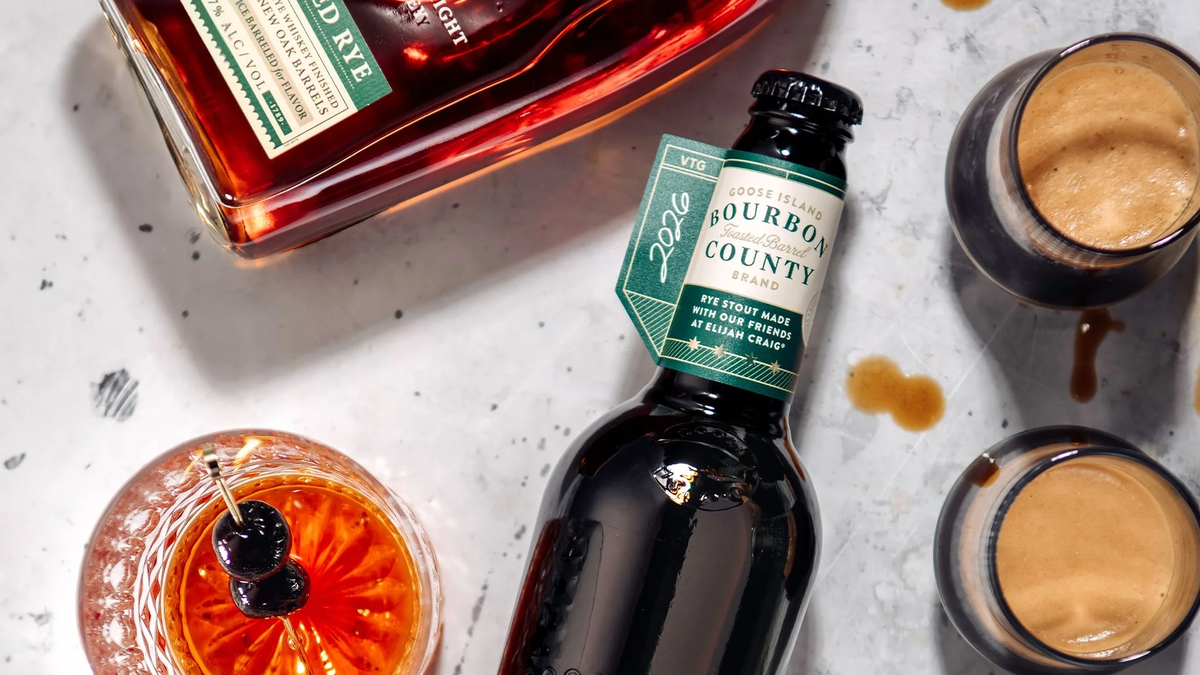 A bottle of 2026 Goose Island Bourbon County Toasted Barrel Rye Stout with a green label laid on a white marbled surface. It is accompanied by a bottle of Elijah Craig Toasted Rye whiskey, two small glasses of stout, and a bright cocktail garnished with cherries on a skewer.