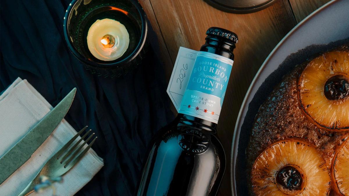 A bottle of 2026 Goose Island Proprietor's Bourbon County Upside-Down Cake Stout featuring a light blue label. It sits next to a baked pineapple upside-down cake on a plate, silverware on a napkin, and a lit candle.
