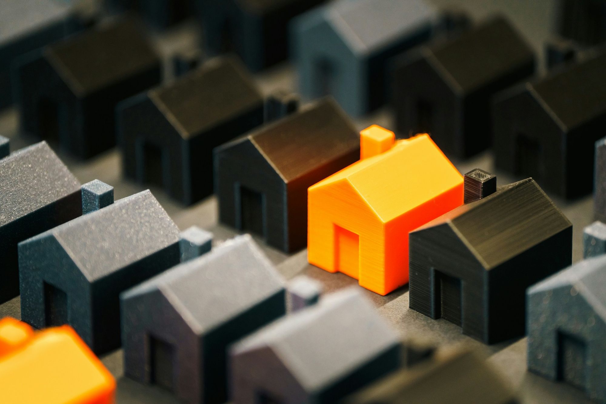 Rows of dark miniature model houses with a single bright orange house standing out