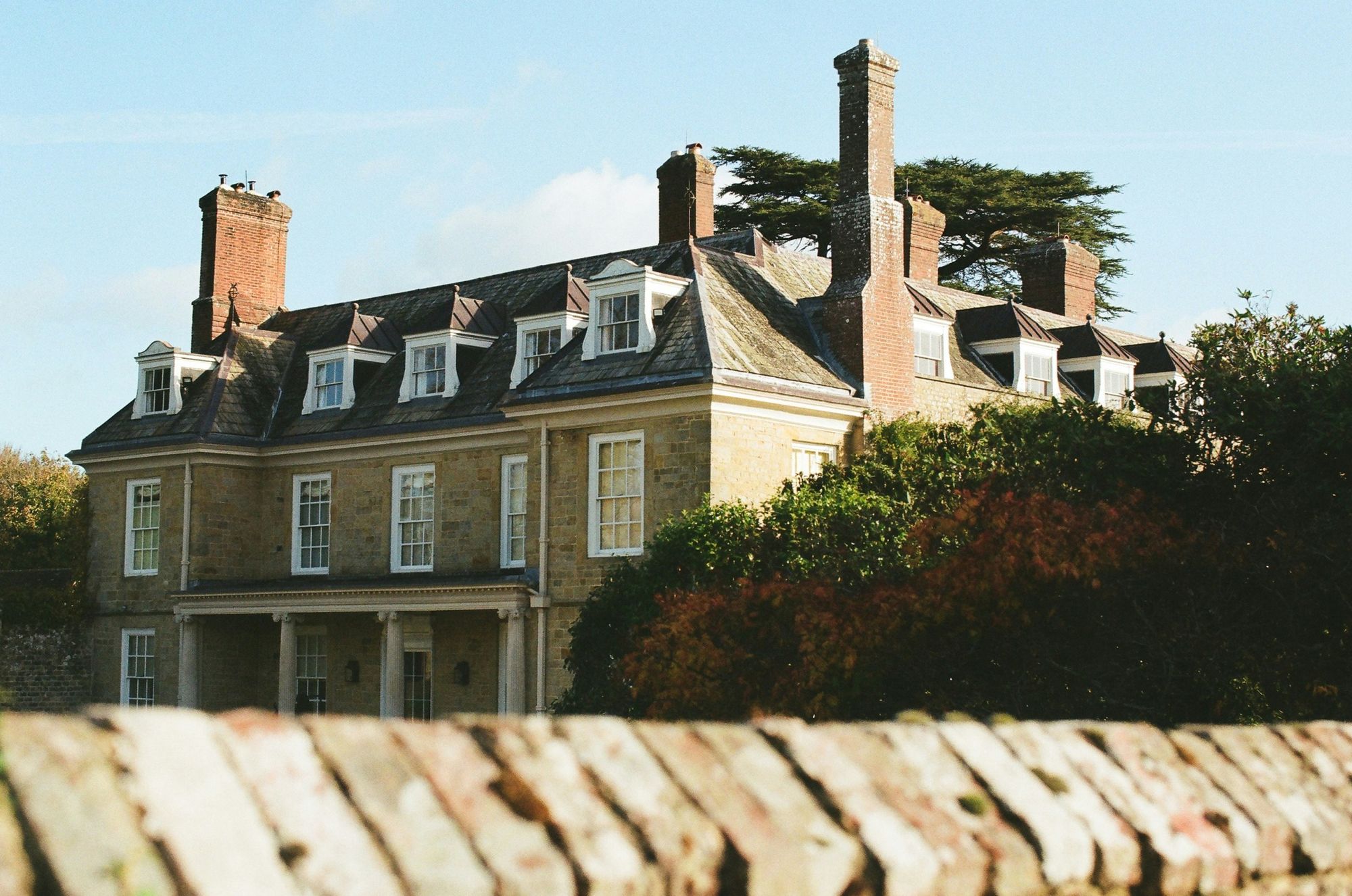 Large stone manor house with tall chimneys and dormer windows behind a low wall