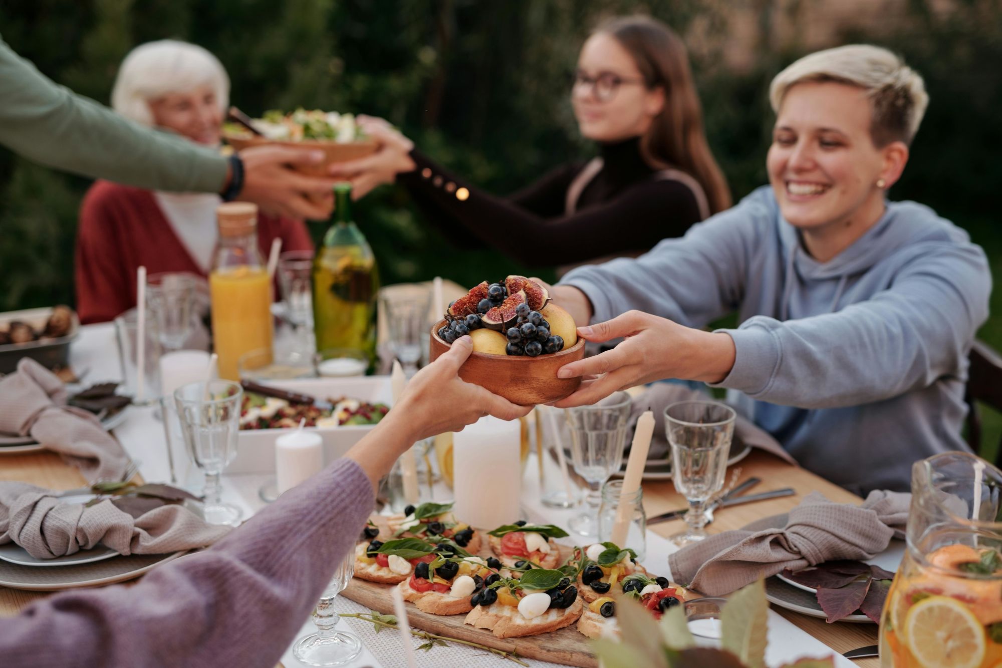 Friends passing bowls of figs and grapes across a candlelit outdoor dinner table