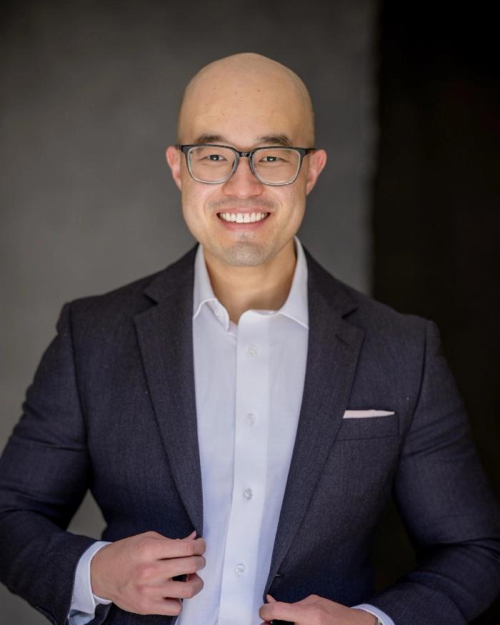 Dontcher Bramlett in glasses and a navy blazer, smiling against a dark backdrop