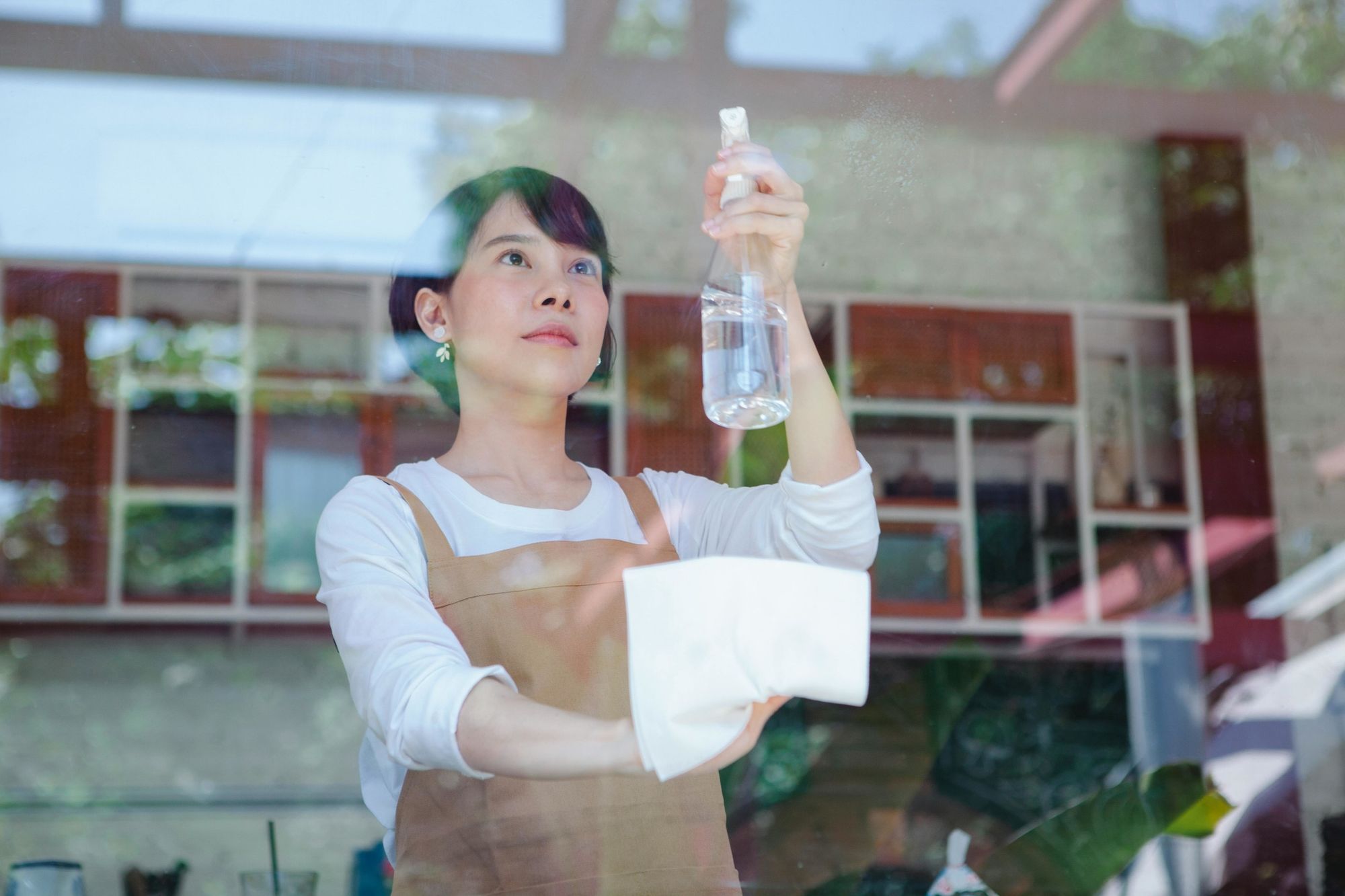 Woman in an apron cleaning a window with a spray bottle and white cloth