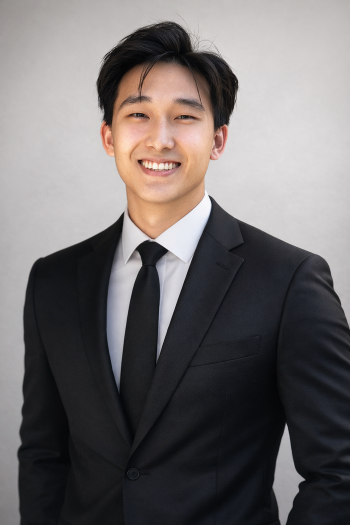 Noah Kim in a black suit and tie, smiling against a light grey wall