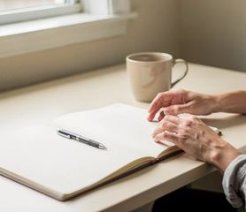 Un quaderno aperto, una penna e una tazza su una scrivania chiara accanto a una finestra.