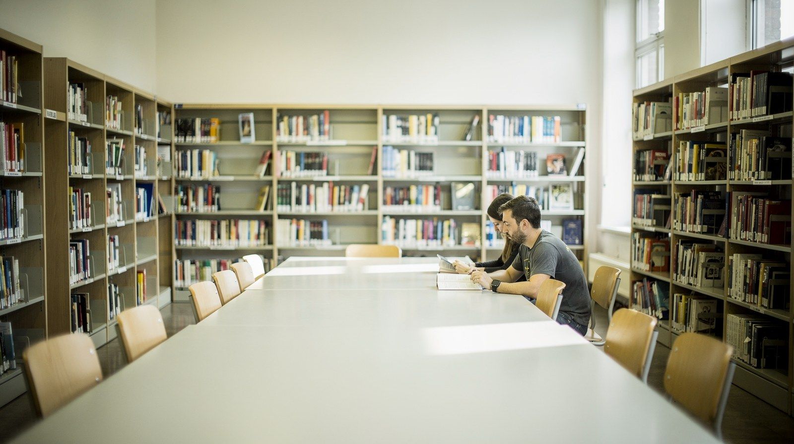 Una sala di lettura luminosa con scaffali alti e un lungo tavolo vuoto.
