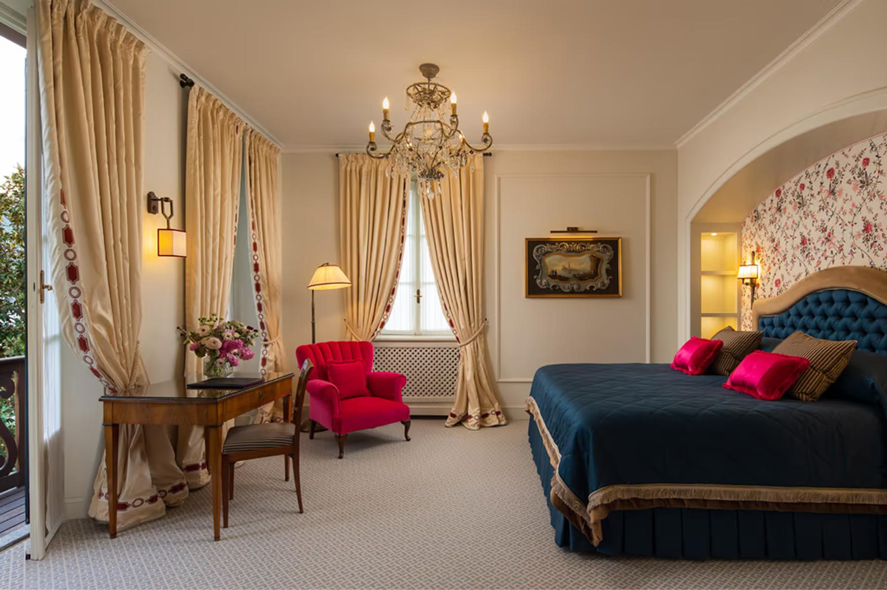 Suite with red velvet armchair, gold bed frame, tall arched window, and frescoed ceiling