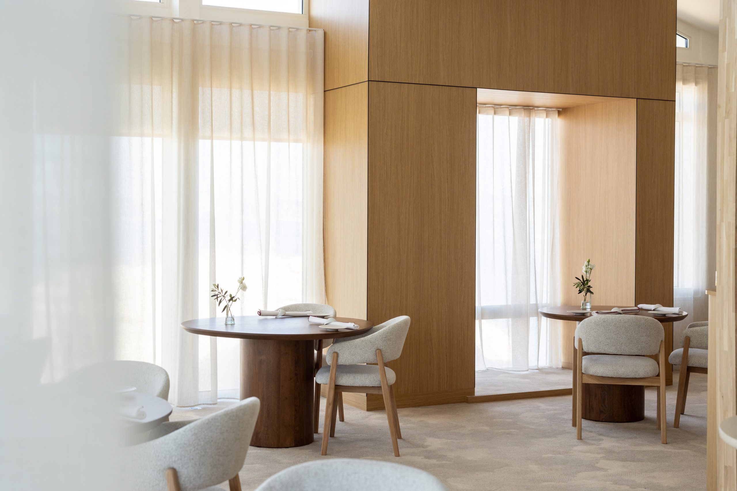 A pale wood dining room, round tables and sheer curtains in afternoon light