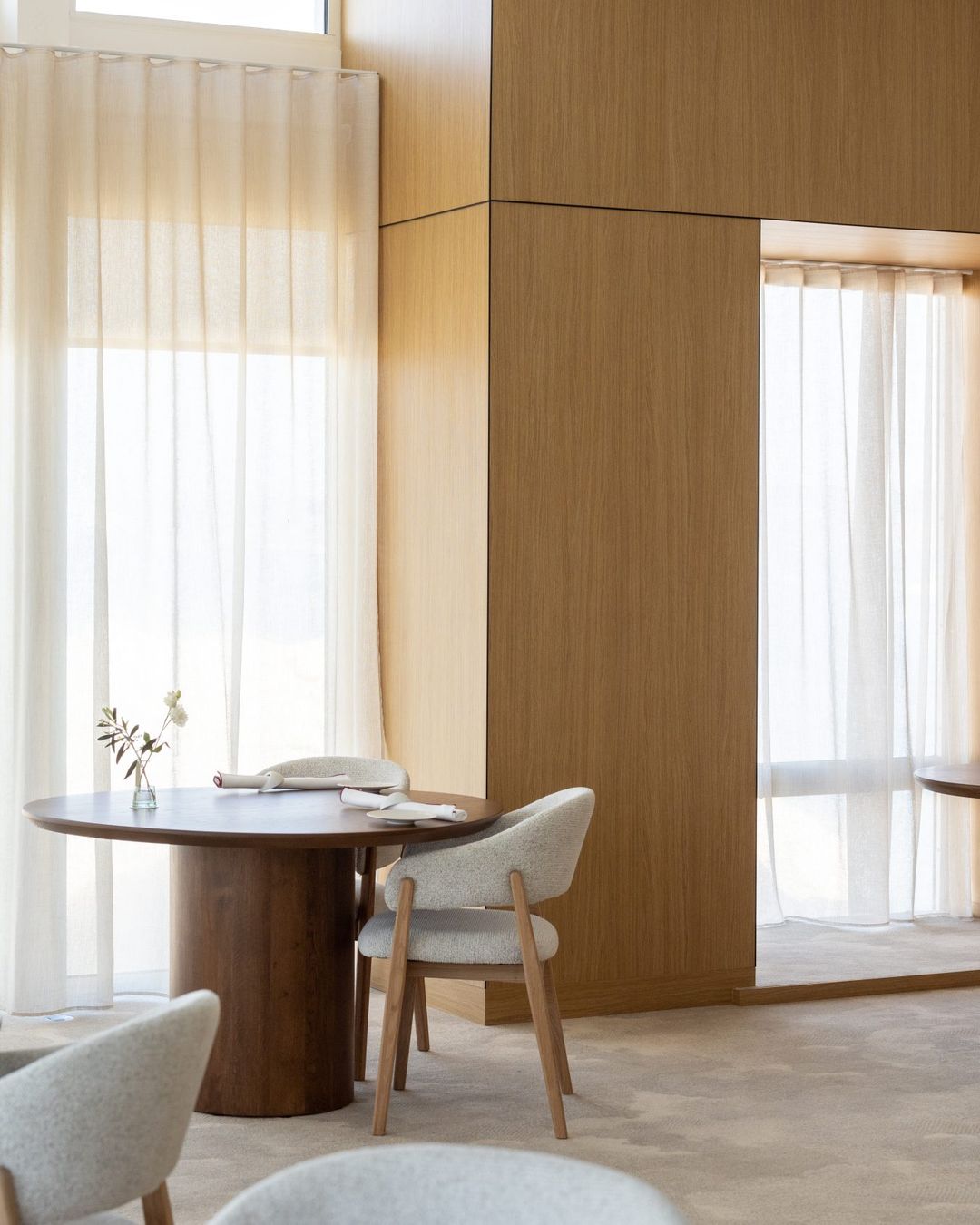 A pale wood dining room, round tables and sheer curtains in afternoon light