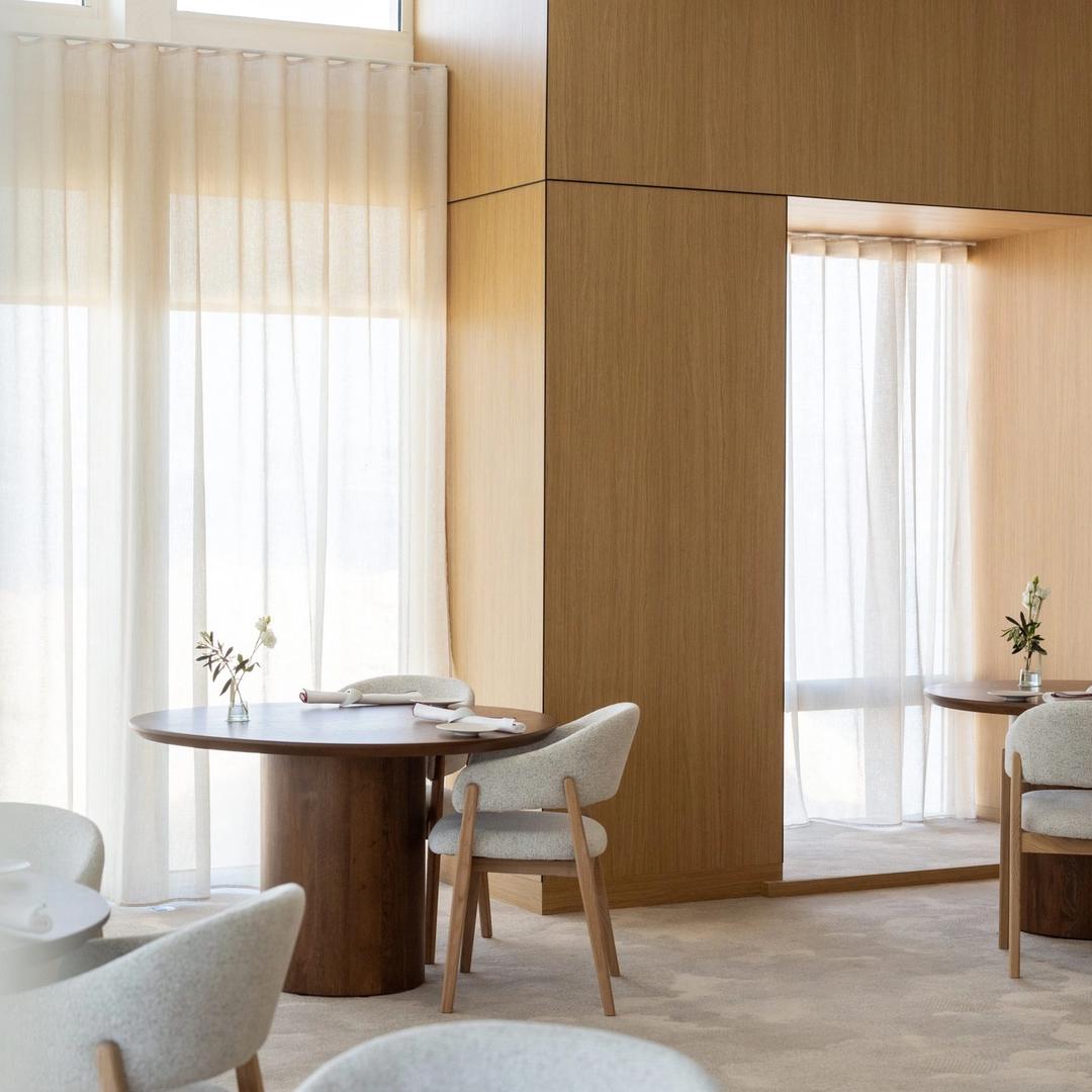 A pale wood dining room, round tables and sheer curtains in afternoon light