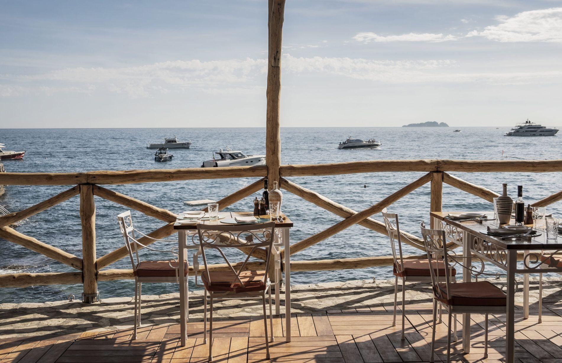 Seaside terrace with white-clothed tables, wooden chairs, and clear Mediterranean water view
