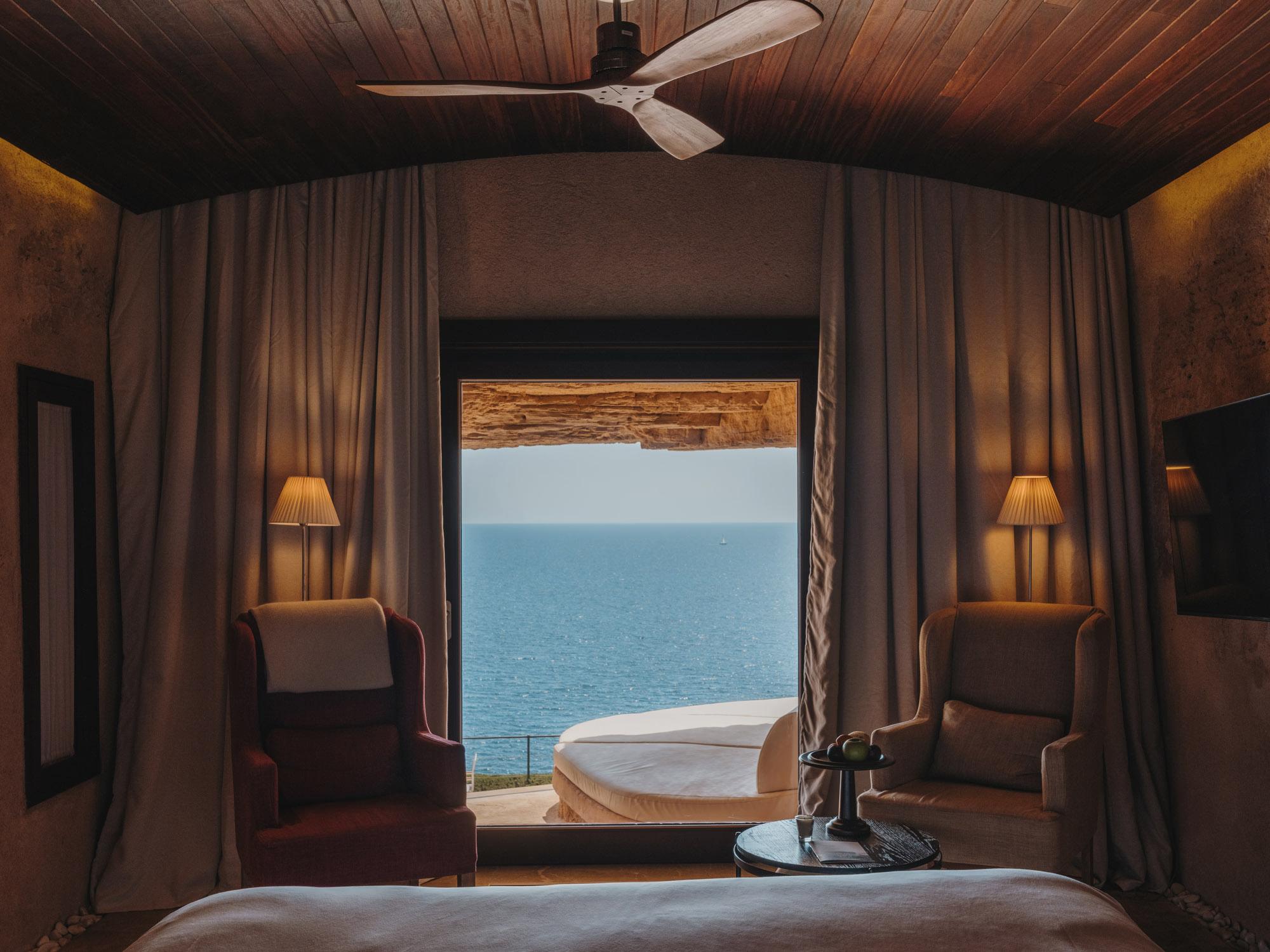 Cap Rocat: a curtained bedroom framing turquoise water through an arched window, with gold sconces and lounge chairs