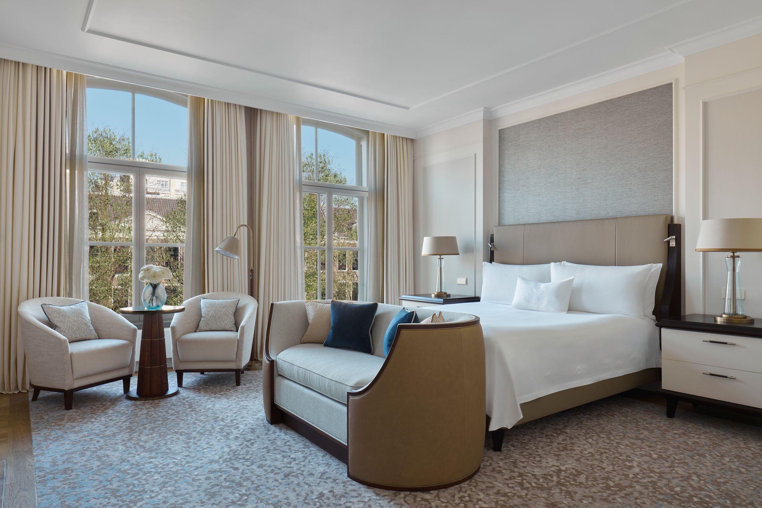Waldorf Astoria Amsterdam: a room with a brown upholstered bed frame, cream seating, and tall arched windows overlooking the canal