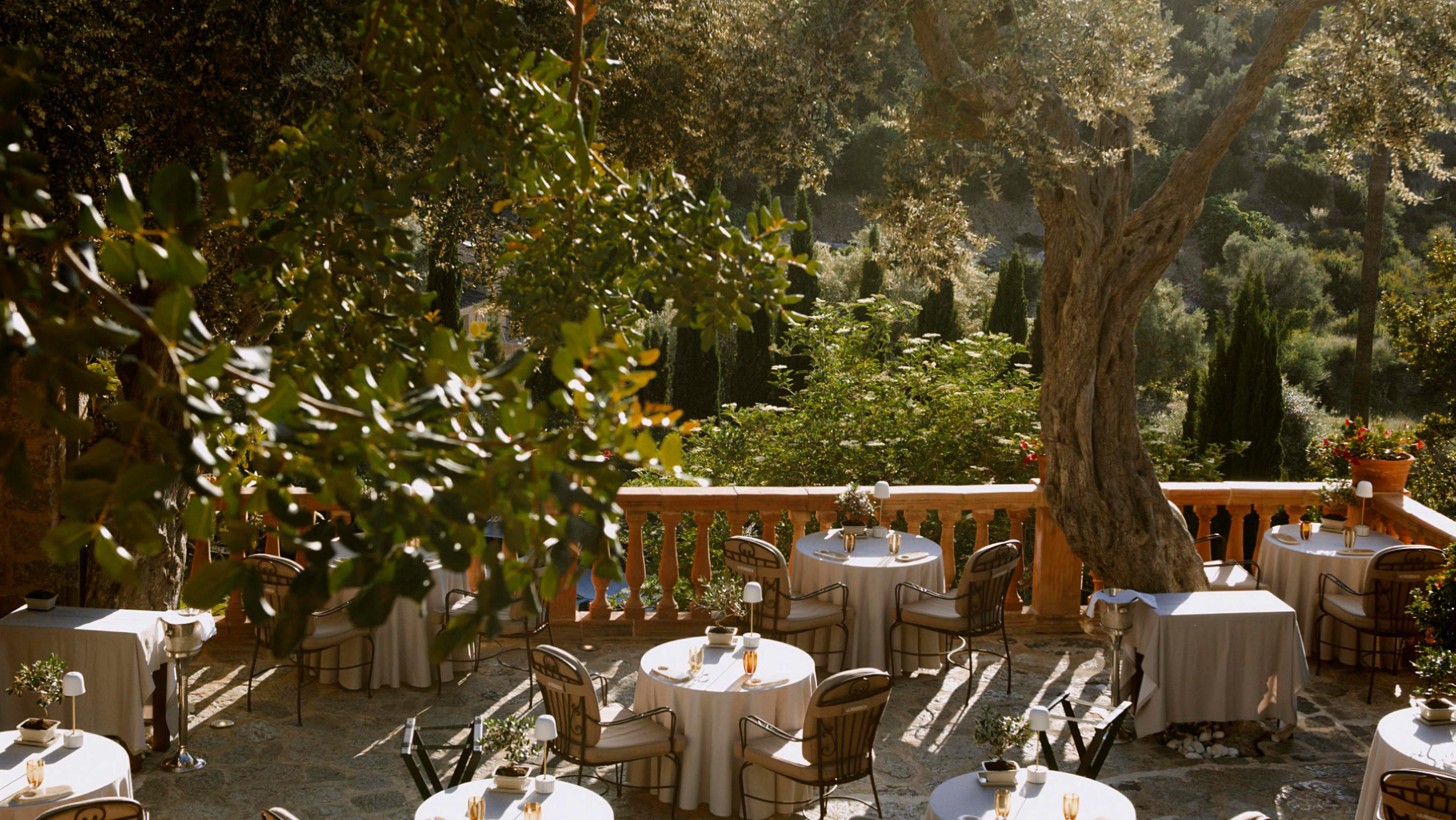 Terraced dining tables with white linens beneath shaded pergolas overlooking Mediterranean foothills