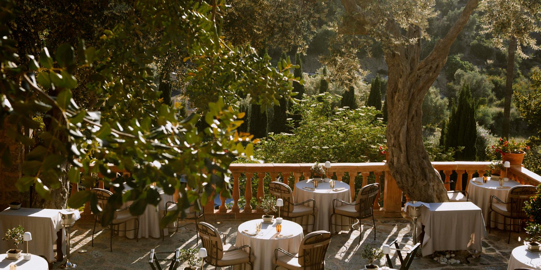 Terraced dining tables with white linens beneath shaded pergolas overlooking Mediterranean foothills