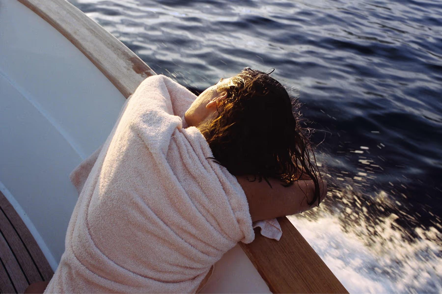 Caruso: person leaning over boat rail gazing at sea with coastal mountains in distance