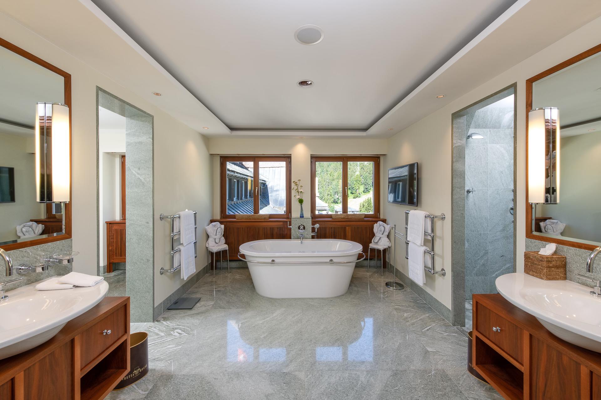 Suvretta House: a spa bathroom with dual sinks, a soaking tub, large windows, and neutral stone finishes
