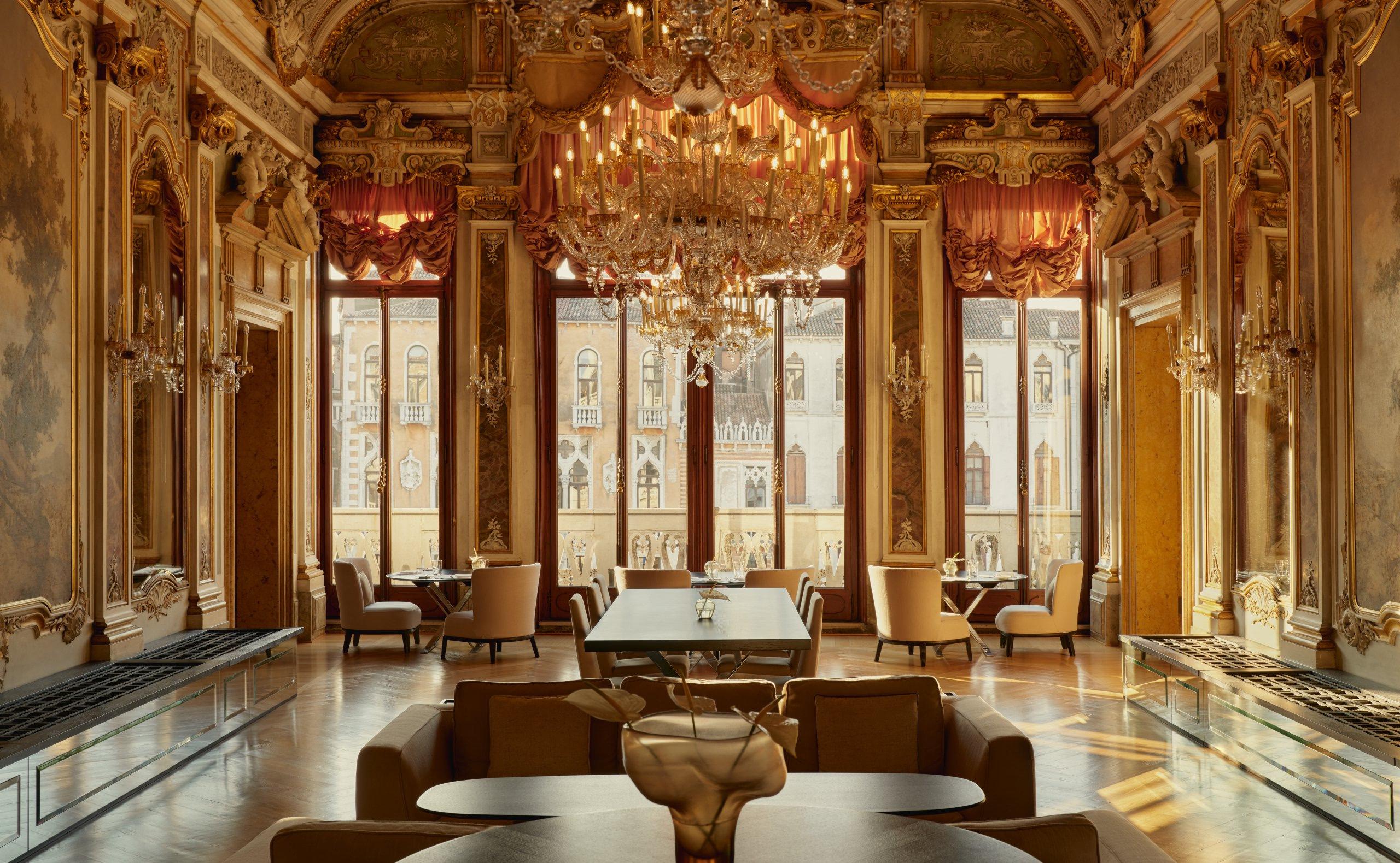 A grand gilded gallery with ornate painted ceiling, crystal chandelier, tall arched windows with columns, and red-toned upholstered seating