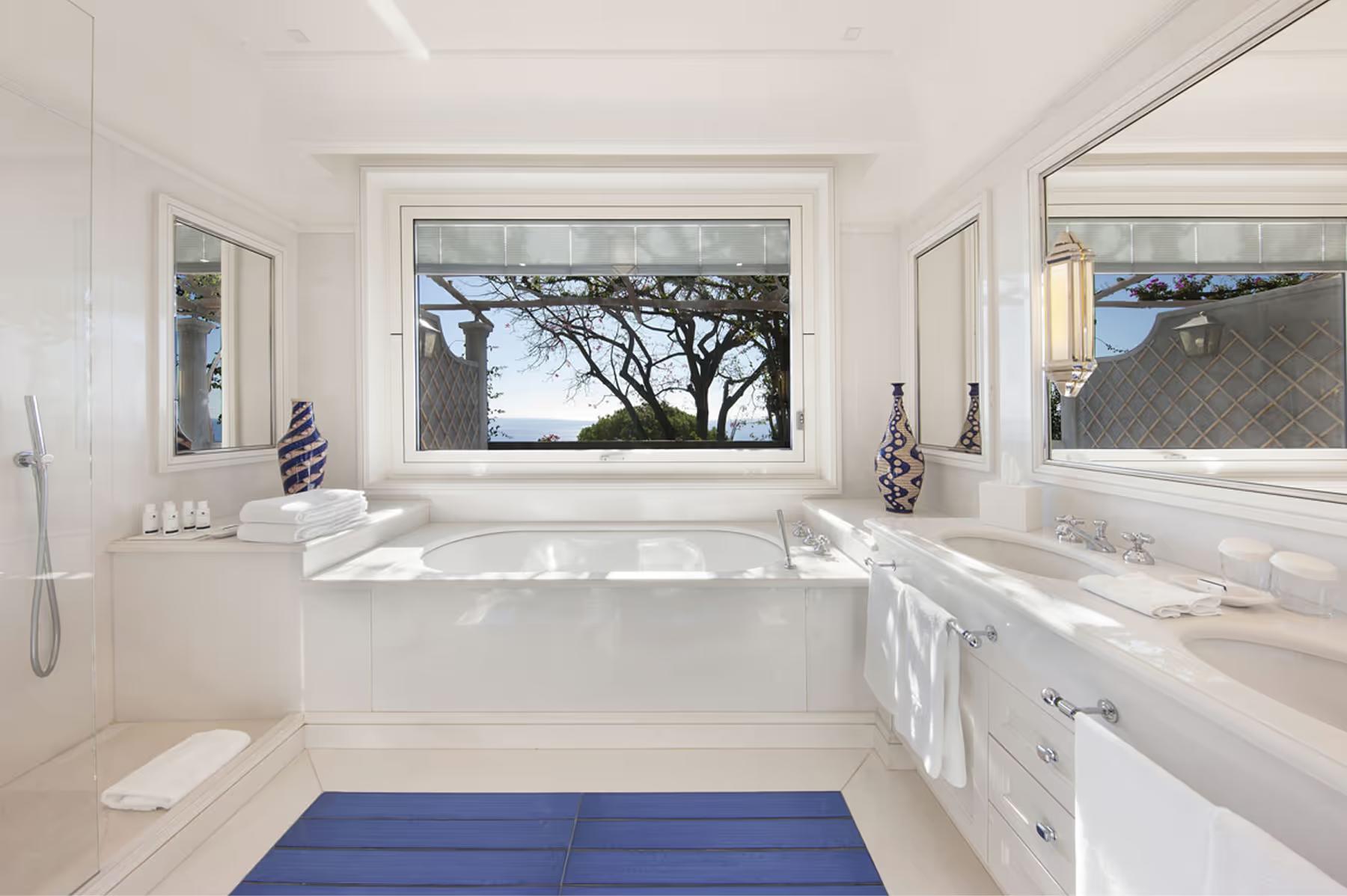 White marble bathroom with view window overlooking Mediterranean landscape, soaking tub, blue accent mat