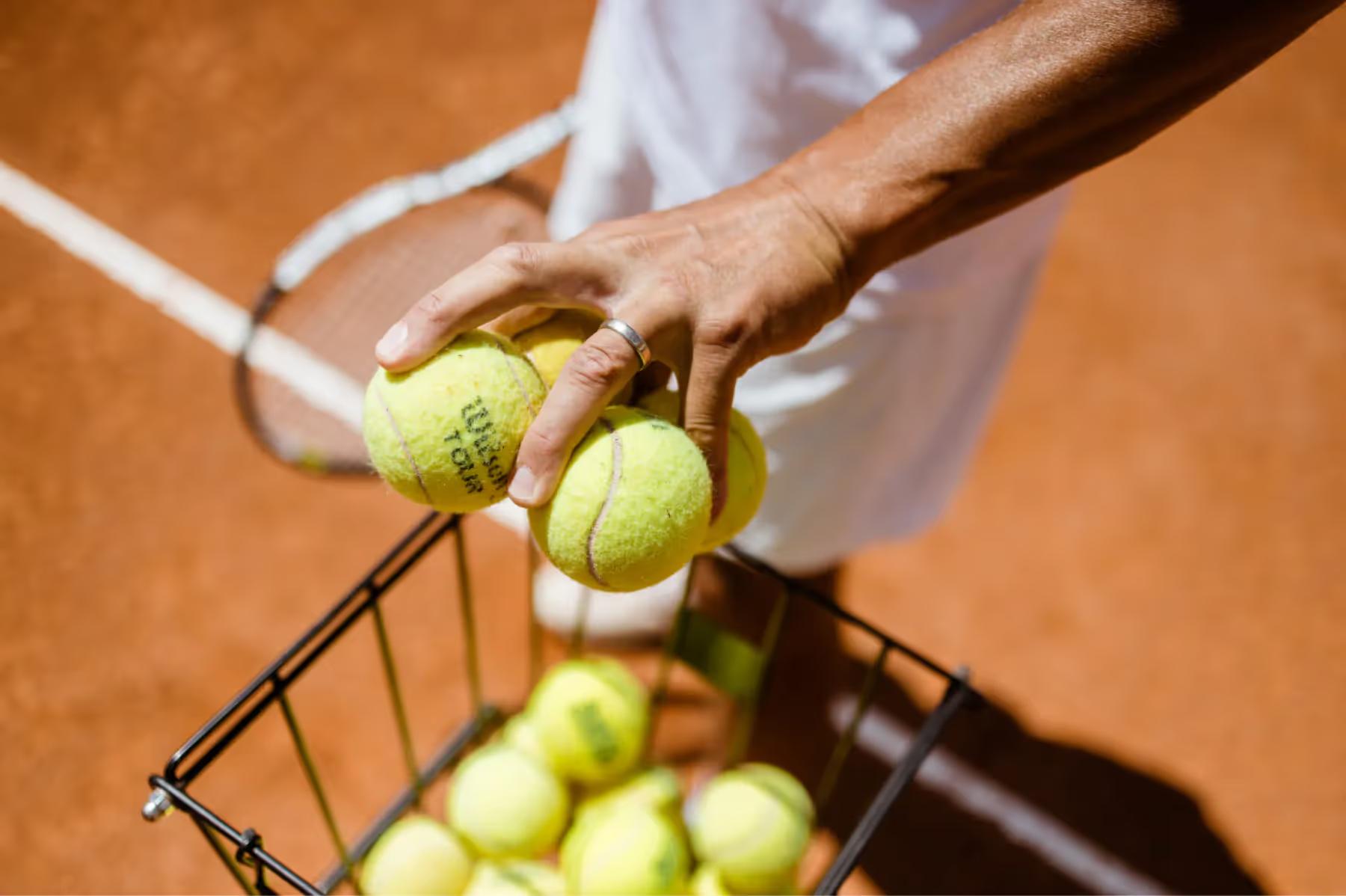 Tennis balls being held above a clay court with a racket and basket of additional balls