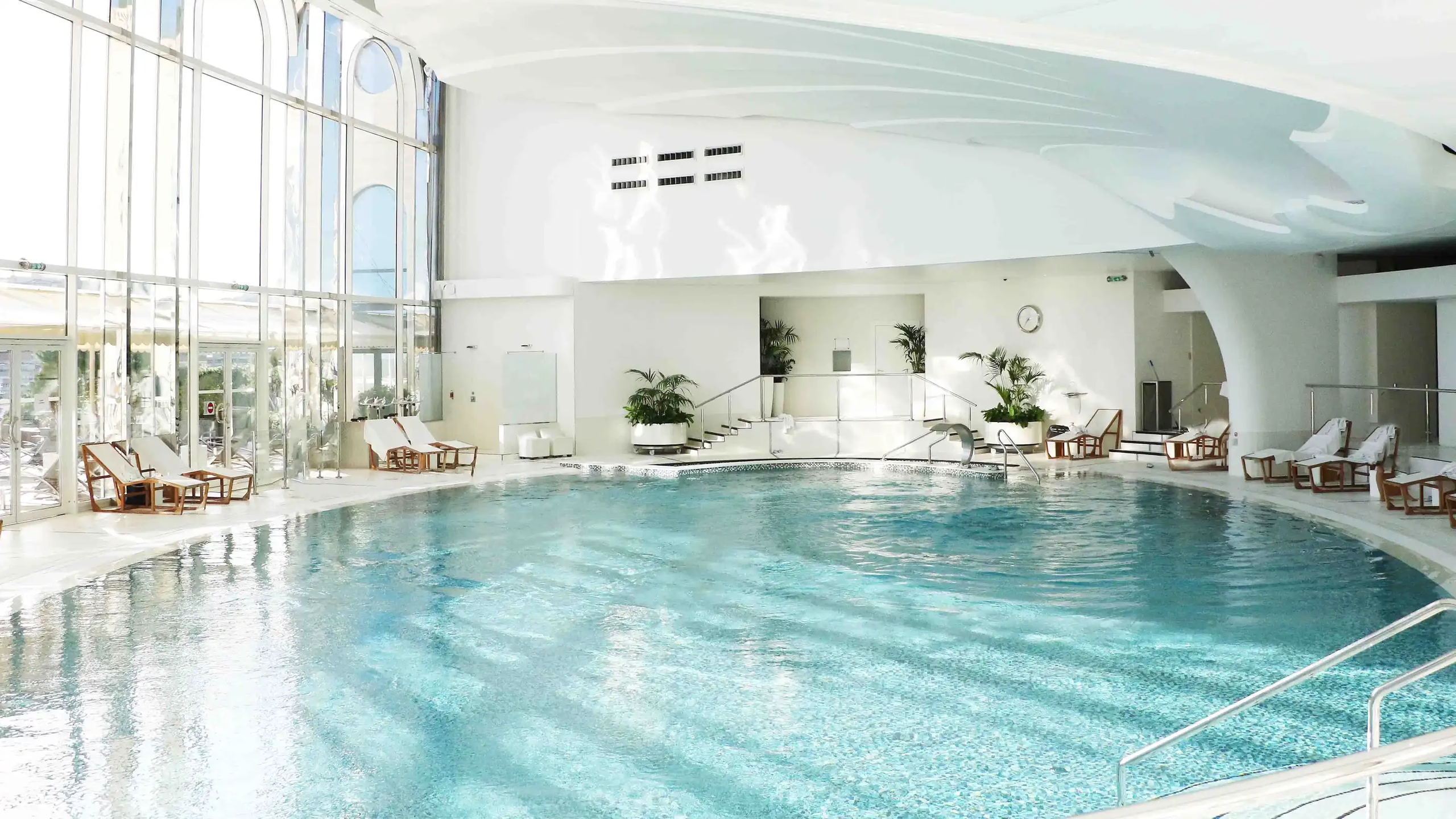 Indoor swimming pool with natural light and rows of lounge seating along deck