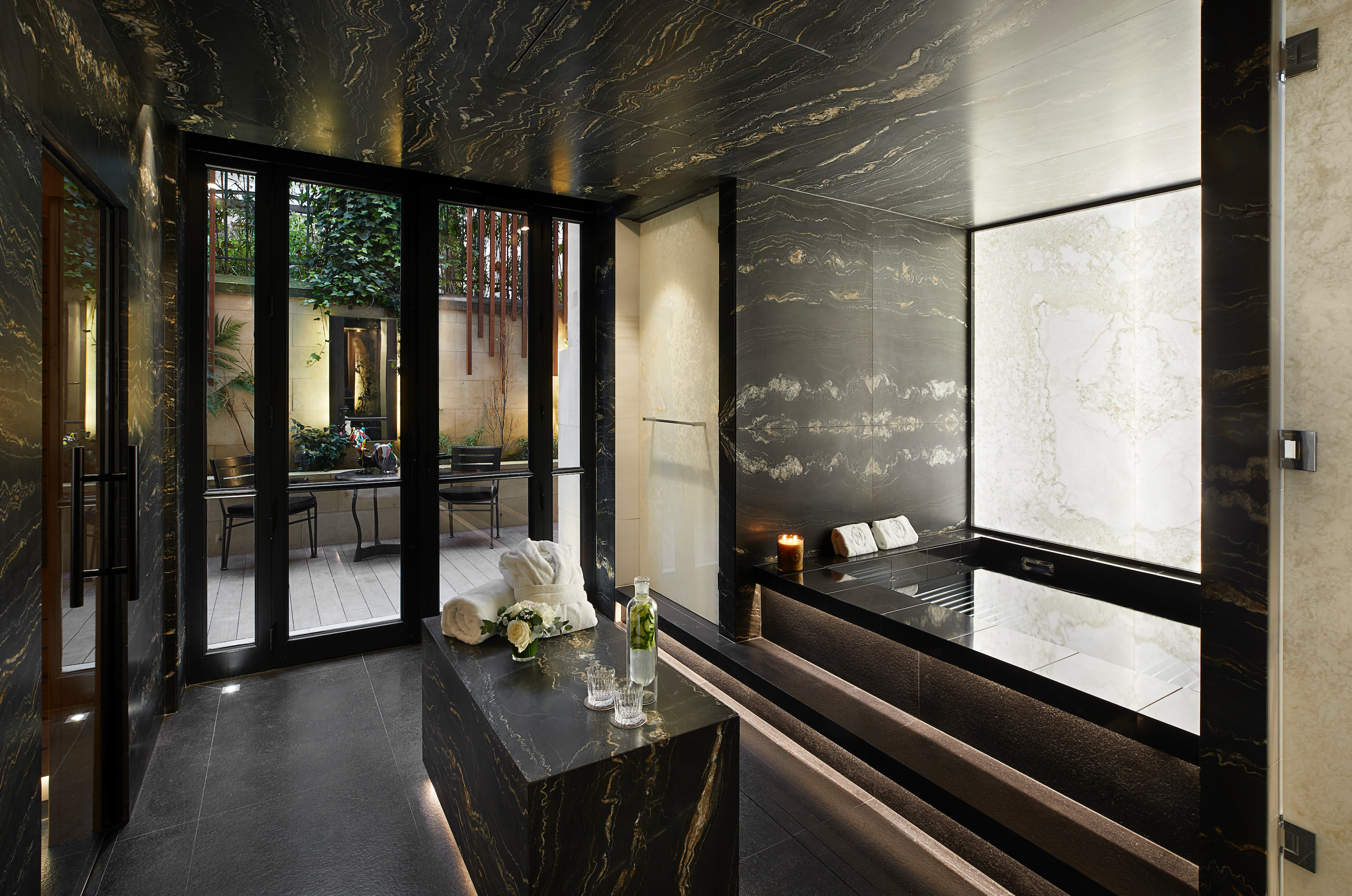 A dark spa interior with glass doors opening to a garden, pale marble walls, and modern furnishings