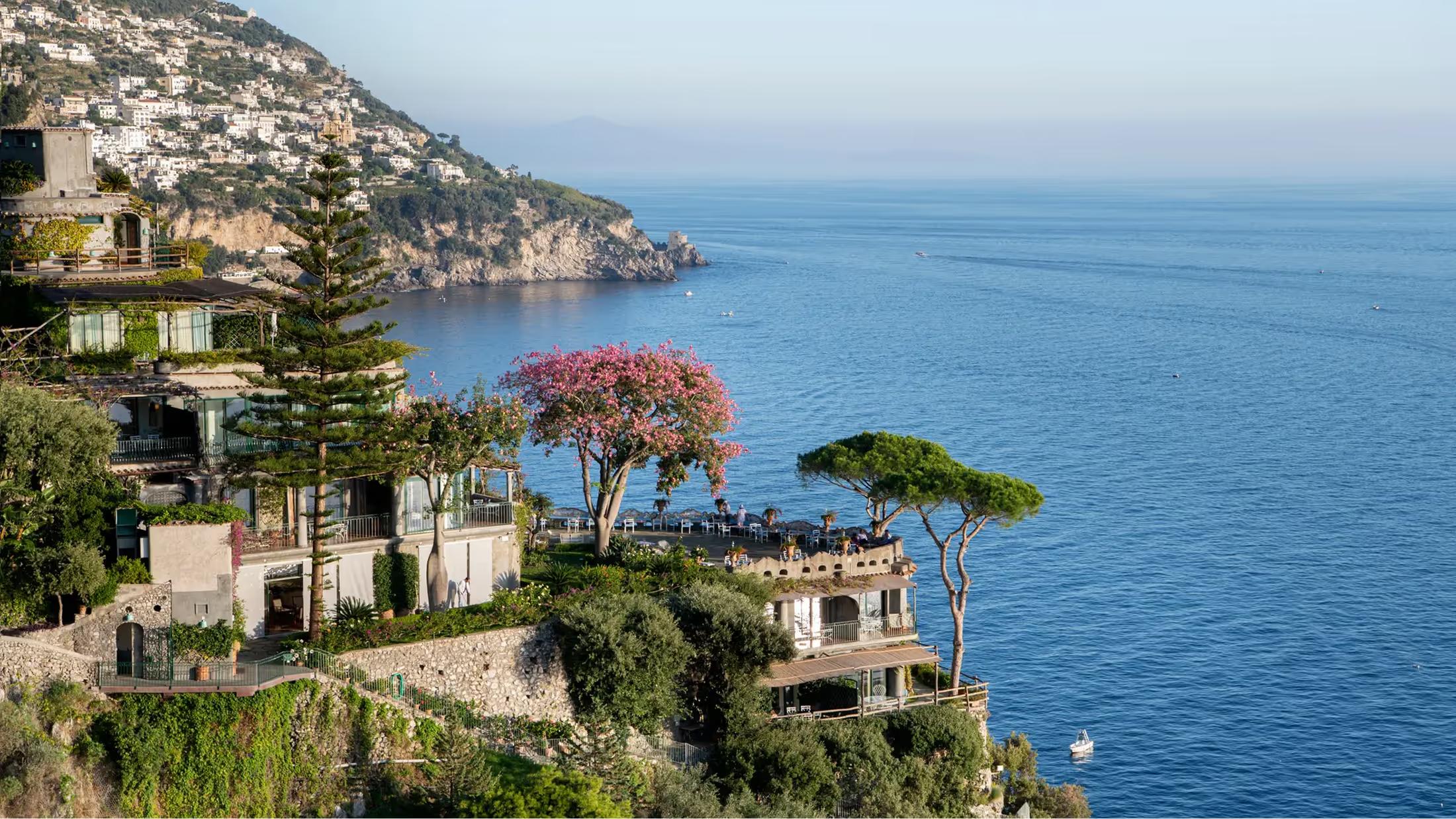 A terraced hotel cliffside above turquoise Mediterranean water with trees and stone buildings