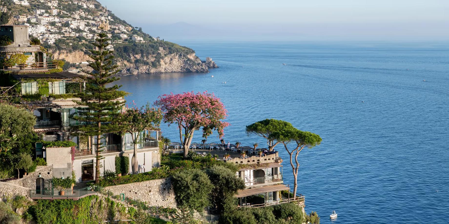 A terraced hotel cliffside above turquoise Mediterranean water with trees and stone buildings